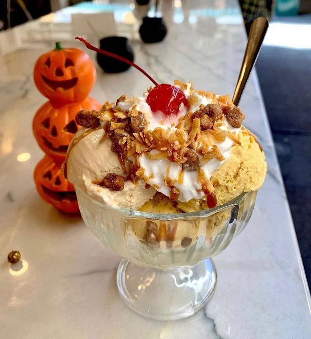 A footed glass sundae with whipped cream and a cherry, small pumpkins beside it.